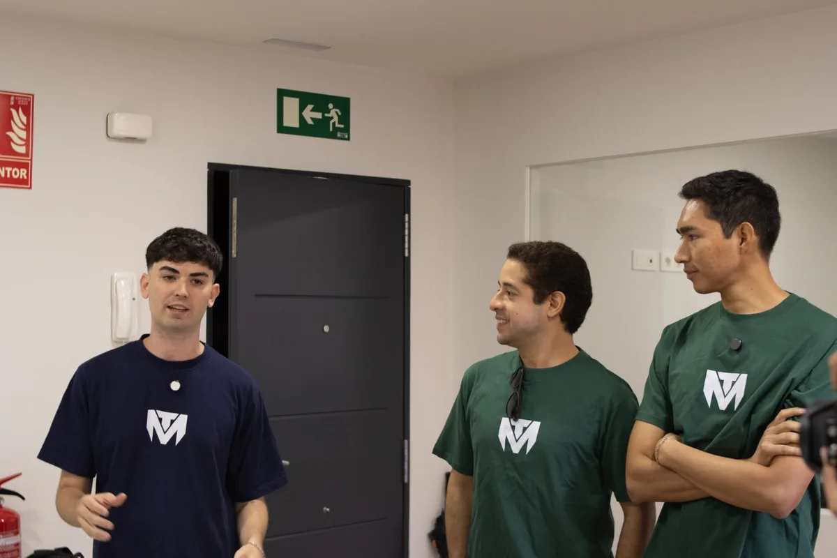 Three men in TuManag3r shirts talk indoors
