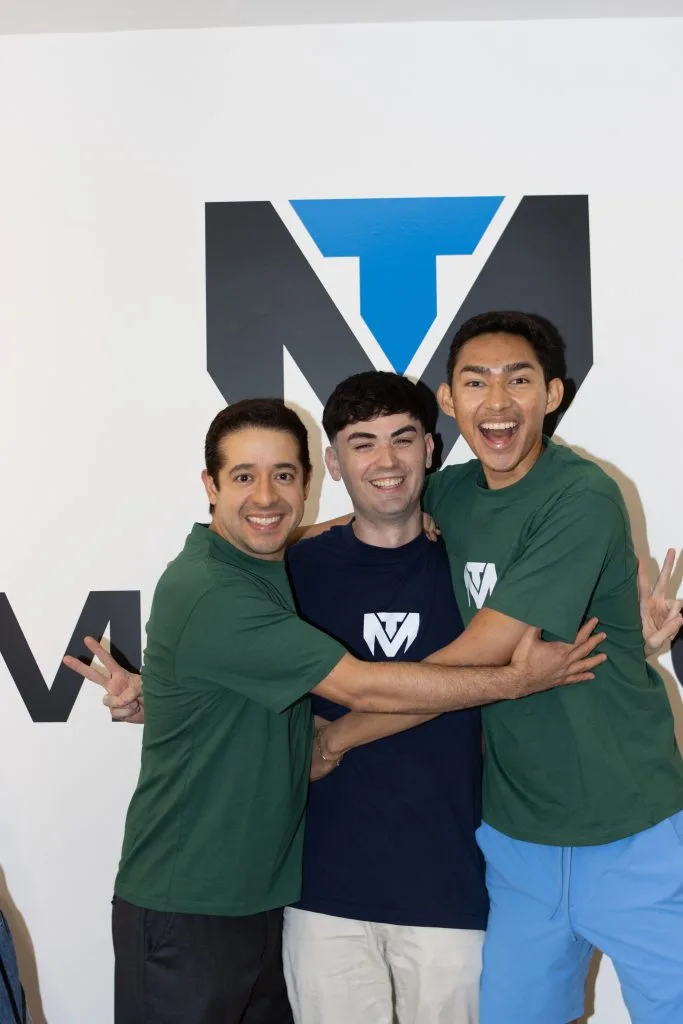 Three men pose in front of the TuManag3r logo